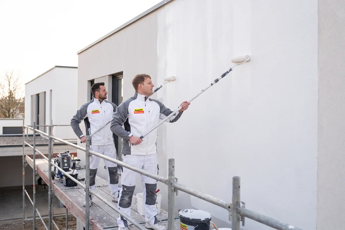 Zwei Maler führen auf einem Gerüst einen Fassadenanstrich an der Außenwand eines modernen Gebäudes durch