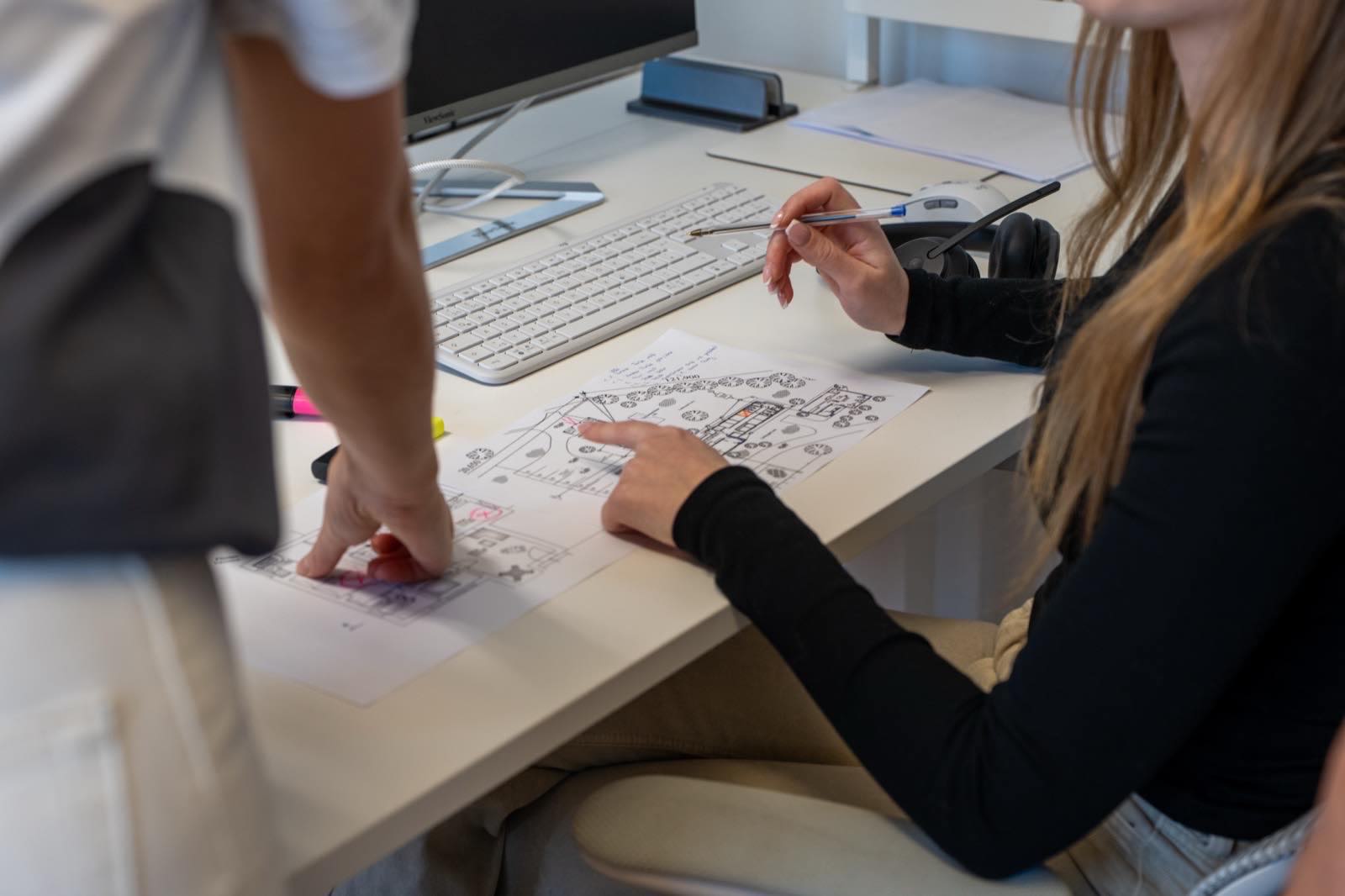 Zwei Personen besprechen aufmerksam Architekturpläne an einem Schreibtisch im Büro mit Tastatur und Monitor
